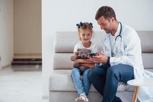 Male doctor in white uniform sits in the clinic with little girl photo