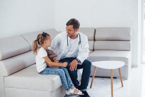 Male doctor in white uniform sits in the clinic with little girl photo