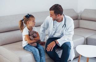 Male doctor in white uniform sits in the clinic with little girl photo