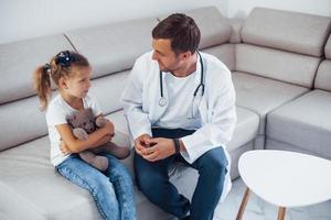 Male doctor in white uniform sits in the clinic with little girl photo