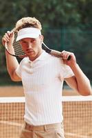 Posing for a camera. Young tennis player in sportive clothes is on the court outdoors photo