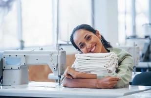 Young positive dressmaker sits by the table with clothes on it photo