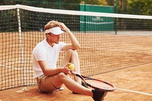 Tired tennis player in sportive clothes is on the court outdoors leaning on the net photo