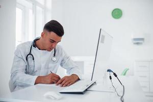 Working with documents. Young stylish male doctor with stethoscope is in clinic at daytime photo