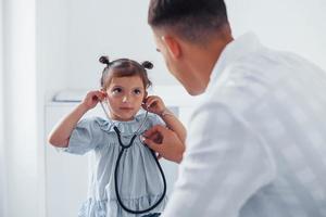 enseña a la niña a usar el estetoscopio. joven pediatra trabaja con una pequeña visitante en la clínica
