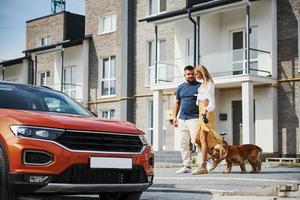 Going out from the building. Lovely couple have a walk together with dog outdoors near the car photo