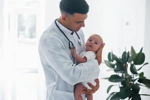 Stands against window. Young pediatrician is with little baby in the clinic at daytime photo