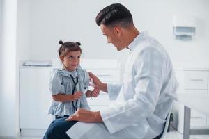 Teaches girl how to use stethoscope. Young pediatrician works with little female visitor in the clinic photo