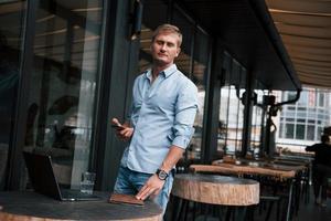 Holds phone in hand. Front view of guy in jeans that stands in the cafe at daytime photo