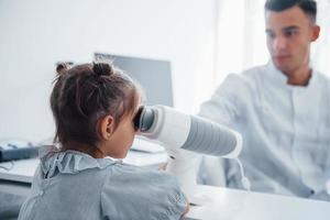Looks into special device. Young ophthalmologist is with little female visitor in the clinic photo