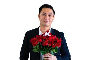 hombre asiático vestido con traje negro sosteniendo un ramo de rosas rojas y una caja de regalo roja aislada en fondo blanco para el concepto de aniversario o día de san valentín. foto