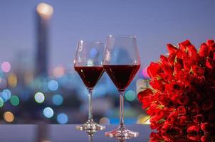 dos copas de vino tinto y ramo de rosas rojas en la mesa con coloridas luces de bokeh de la ciudad para el concepto de aniversario o día de san valentín. foto