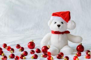 Selective focus on Santa claus teddy bear eyes who wearing hat sitting with red Christmas baubles on white cloth background. photo