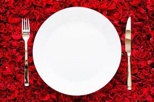 White plate with knife and fork on red rose background for dining, anniversary and Valentine day concept. photo