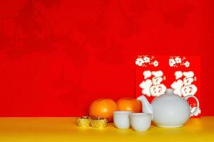 Chinese tea set put with ingots, oranges and red envelope packets or ang bao word mean wealth with shadow of peach blossom on yellow and red glitter paper background.