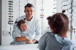 Young pediatrician in white coat helps to get new glasses for little girl photo