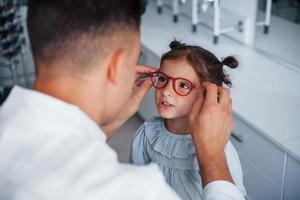 Young pediatrician in white coat helps to get new glasses for little girl photo