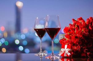 dos copas de vino tinto y un ramo de rosas rojas con una caja de regalo roja en la mesa con coloridas luces de ciudad para el concepto de aniversario o día de san valentín. foto