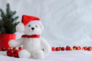 Selective focus on Santa claus teddy bear eyes who wearing hat sitting with red baubles and Christmas tree on white cloth background.
