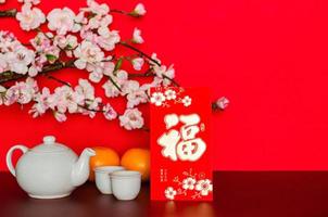 Red envelope packet or ang bao word mean wealth, sentence mean may all go well, great fortune and profit puts with tea set and oranges that have peach blossom on red glitter paper background. photo