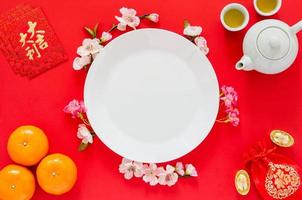 White plate on red background with tea set, gold ingots, red bag word means wealth, ornages, red envelope packets or ang bao word means auspice and chinese blossom flowers for Chinese new year. photo