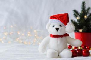 Selective focus on Santa claus teddy bear eyes who wearing hat sitting with red baubles and Christmas tree on white cloth background with lights. photo