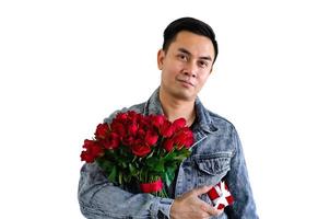 Asian man wearing jeans jacket holding a bouquet of red roses and red gift box isolated in white background for anniversary or Valentine day concept. photo