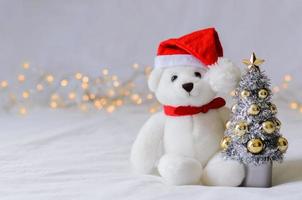 Selective focus on Santa claus teddy bear eyes who wearing hat sitting with blurred focus Christmas tree on white cloth background with lights. photo