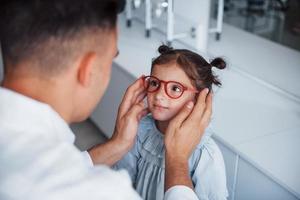 Young pediatrician in white coat helps to get new glasses for little girl photo