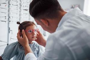 Young pediatrician in white coat helps to get new glasses for little girl photo