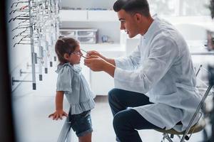 Young pediatrician in white coat helps to get new glasses for little girl photo