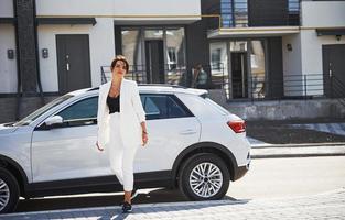 Confident woman in white formal wear outdoots near her white car photo