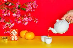 Hand pouring tea from pot for chinese new year concept with oranges, red envelope or ang bao word mean great fortune and ingots on yellow and red glitter paper background. photo