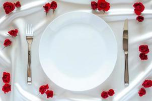 plato blanco con cuchillo y tenedor sobre tela satinada blanca y fondo de rosas rojas para el concepto de cena, aniversario y día de san valentín. foto