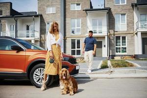 Going out from the building. Lovely couple have a walk together with dog outdoors near the car photo