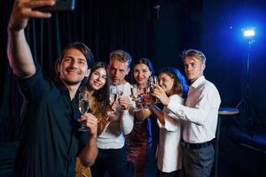 Takes selfie. Group of cheerful friends celebrating new year indoors with drinks in hands photo