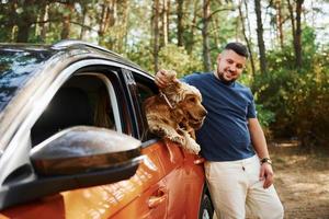 Man stands by the car in the forest. Cute dog in vehicle looks through the window photo