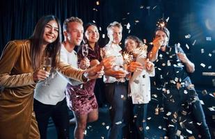 Having fun with sparklers. Confetti is in the air. Group of cheerful friends celebrating new year indoors with drinks in hands photo