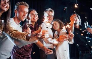 divirtiéndose con bengalas. el confeti está en el aire. grupo de amigos alegres celebrando el año nuevo en el interior con bebidas en las manos foto