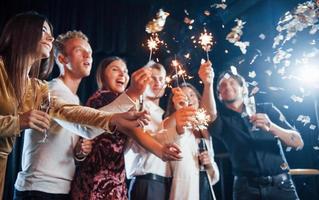 Having fun with sparklers. Confetti is in the air. Group of cheerful friends celebrating new year indoors with drinks in hands photo