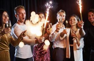 Having fun with sparklers. Group of cheerful friends celebrating new year indoors with drinks in hands