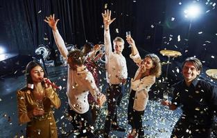 el confeti está en el aire. grupo de amigos alegres celebrando el año nuevo en el interior con bebidas en las manos foto