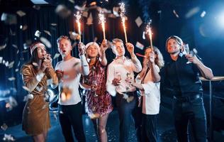 Having fun with sparklers. Group of cheerful friends celebrating new year indoors with drinks in hands photo