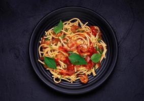 Delicious spaghetti pasta with prawns and cheese served on a black plate. With vegetables, Italian tomato sauce, and spices arranged on a wooden table, black background, top view photo