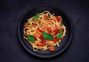 Delicious spaghetti pasta with prawns and cheese served on a black plate. With vegetables, Italian tomato sauce, and spices arranged on a wooden table, black background, top view photo