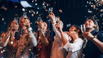 el confeti está en el aire. grupo de amigos alegres celebrando el año nuevo en el interior con bebidas en las manos foto