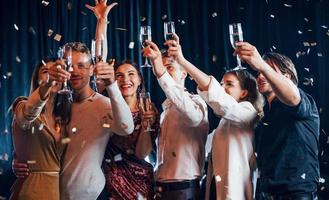 Confetti is in the air. Group of cheerful friends celebrating new year indoors with drinks in hands photo