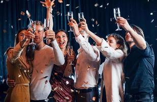 el confeti está en el aire. grupo de amigos alegres celebrando el año nuevo en el interior con bebidas en las manos foto