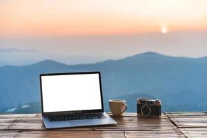 portátil de pantalla vacía con taza de café y cámara con vista a la montaña al amanecer en la mañana foto