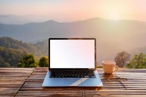 Empty screen laptop with cup of coffee and camera with mountain view at sunrise in the morning photo